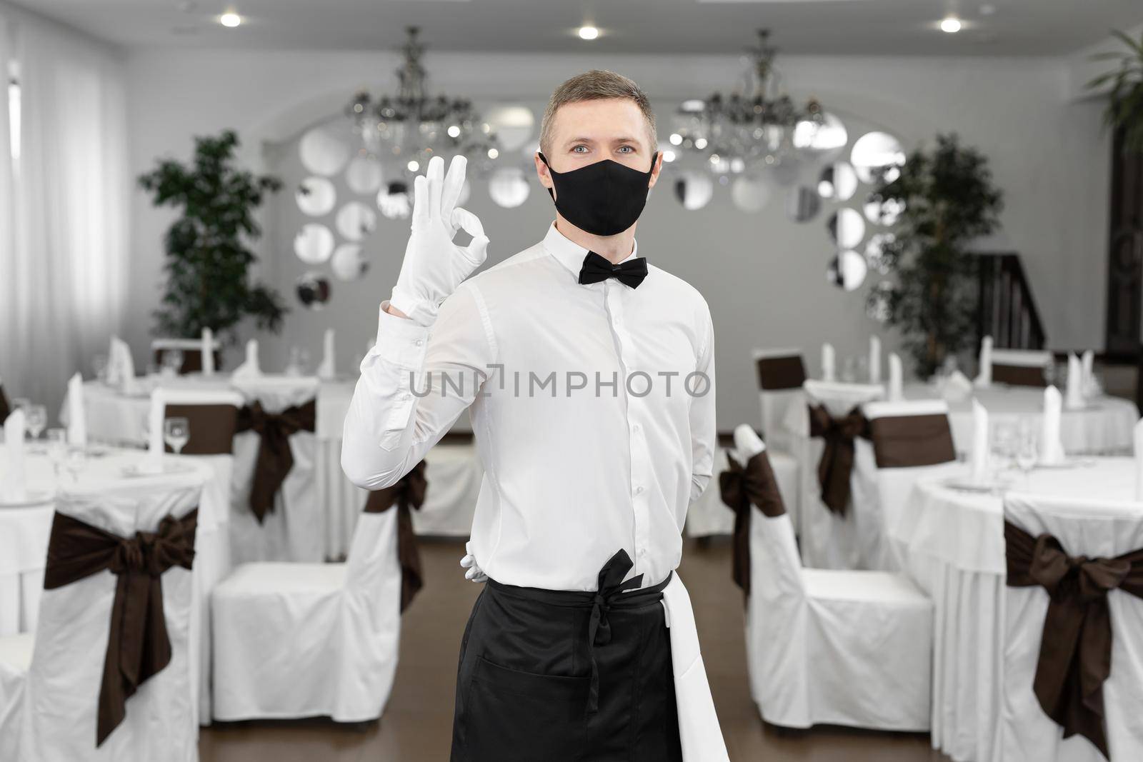 A male waiter in a white shirt and a protective medical mask in the restaurant hall shows the ok sign.