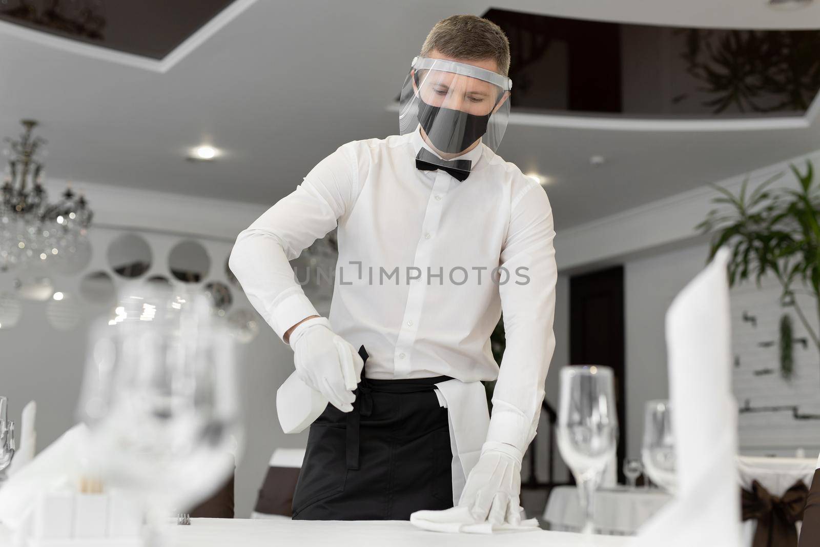 Portrait of a male waiter wearing a face mask and face shield, cleaning a table in a cafe during social distancing. by StudioPeace