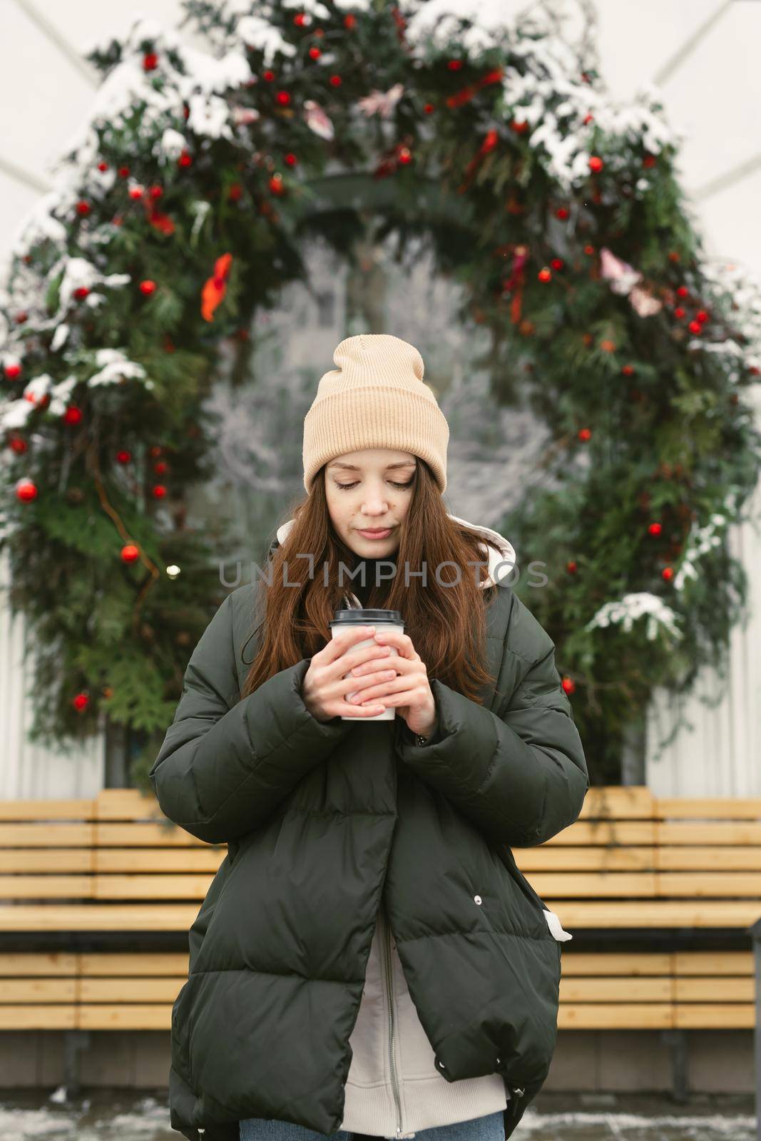 Woman drinks a hot drink on a city street against the background of a cafe window, which is decorated with a Christmas wreath by StudioPeace