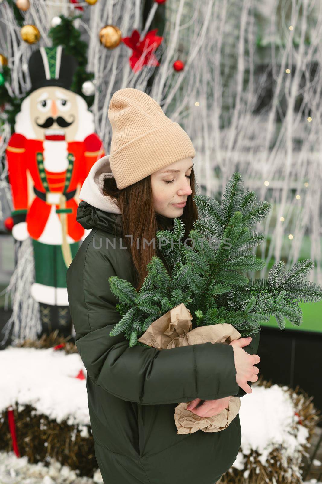 Young woman with a bouquet of Christmas tree branches in a craft package in her hands in winter near the cafe's window by StudioPeace