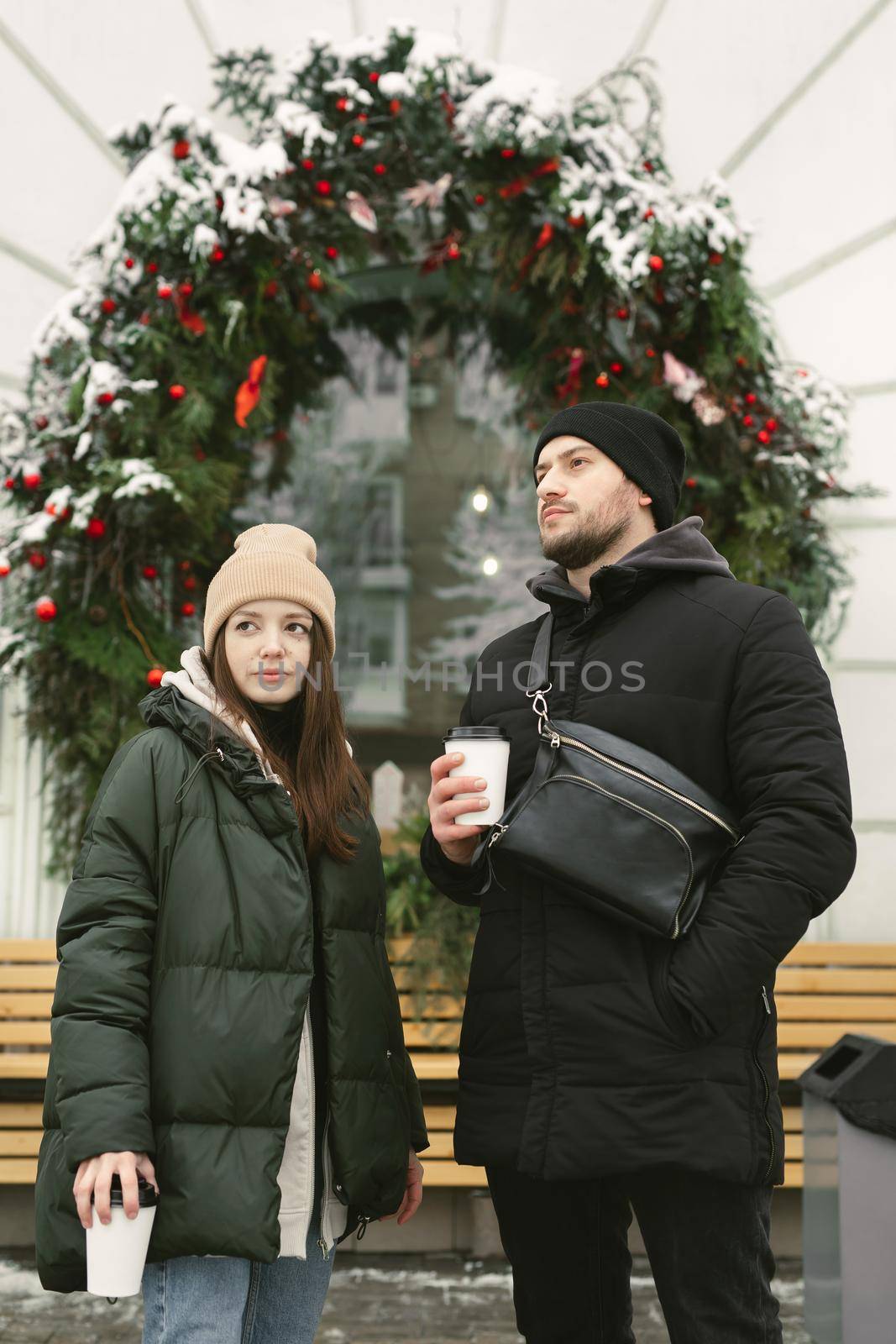 Portrait of a couple on a winter day. A man and a woman are drinking coffee on the street near the decorated window of a cafe by StudioPeace
