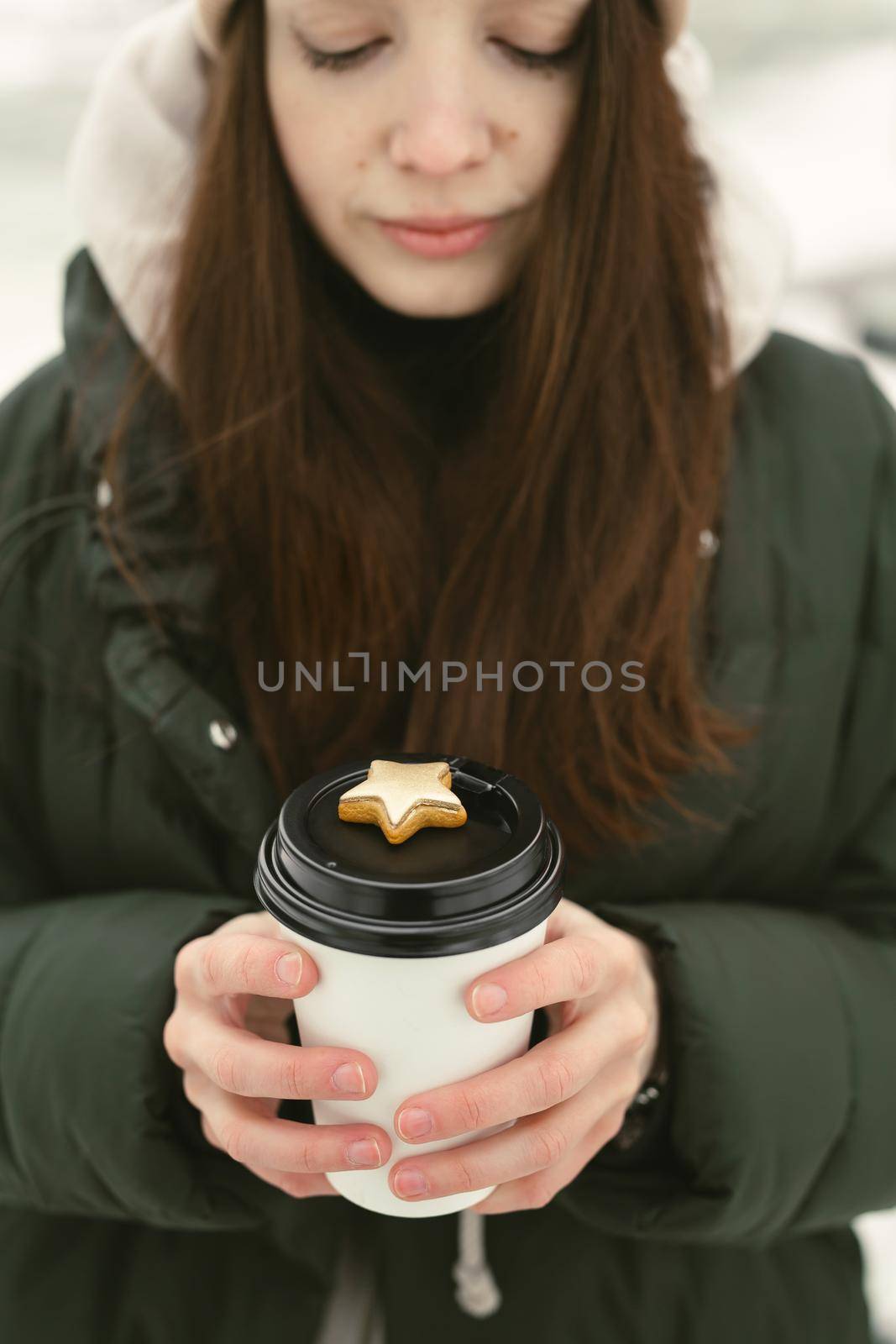 Smiling young woman is holding a mug of hot tea or coffee with gingerbread on the background of a snow-covered garden.