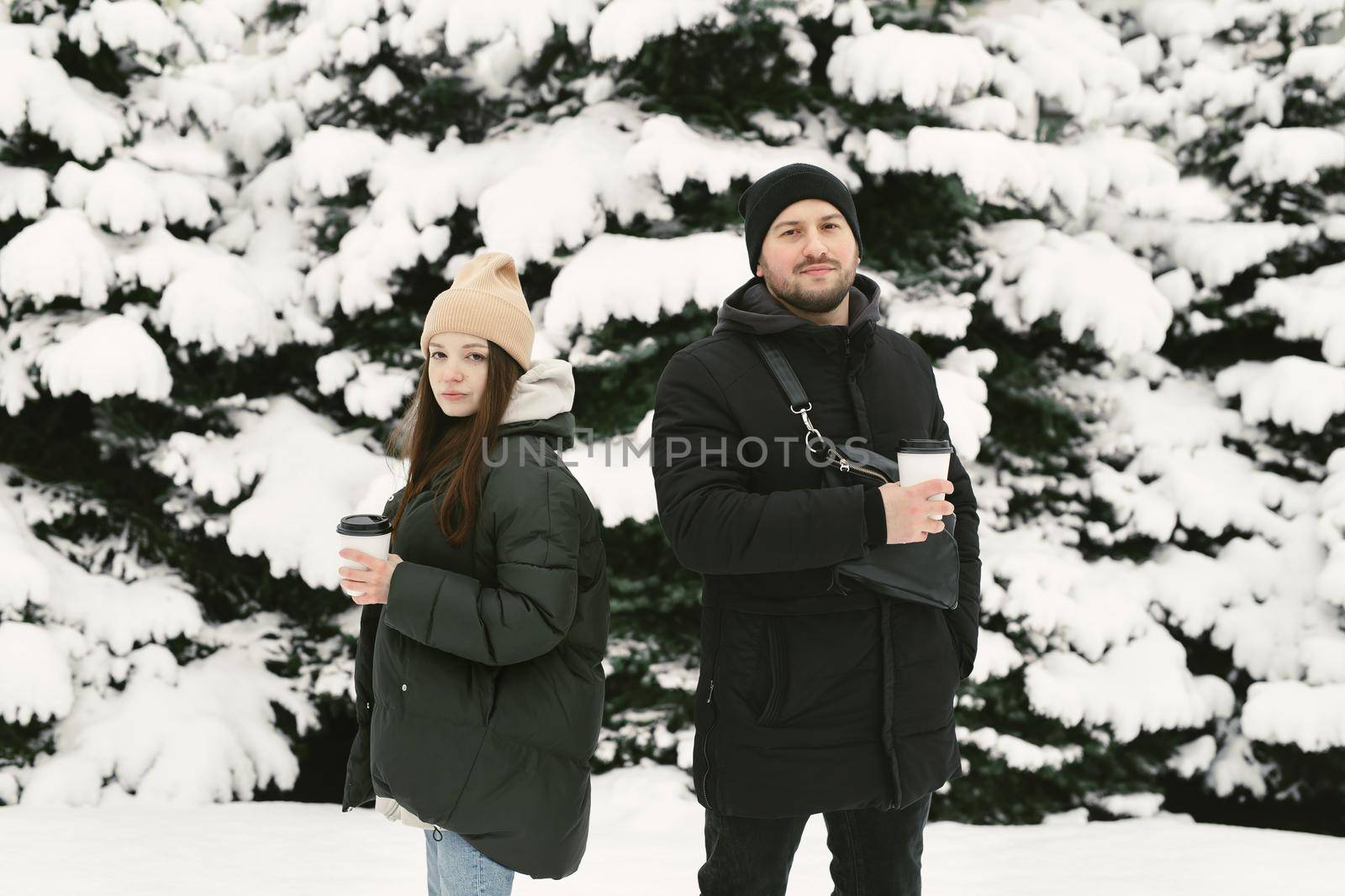 The man and the woman have a rest in the winter park. Young friends in winter style clothes standing among snowy trees and enjoying first snow.