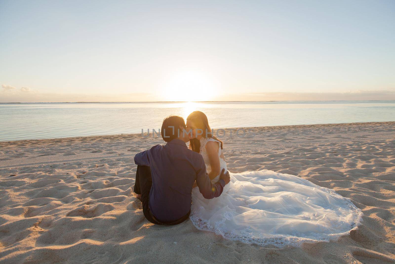 bride and groom sit on the sand. by StudioPeace