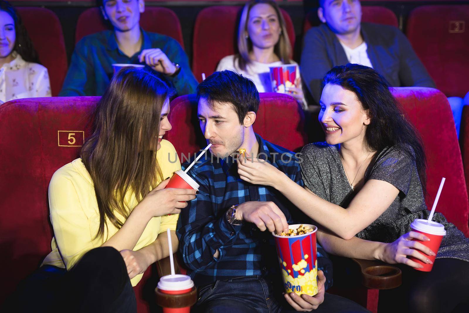 Two young girls and a guy watching a comedy in a cinema. Young friends watching movie in cinema. Group of people in theater with popcorn and drinks. by StudioPeace