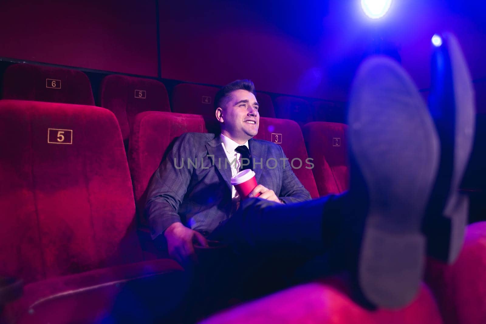 Portrait of a young, handsome man alone in a movie theater in a business suit, with his feet on the front seat and drinking through a tube from a red Cup.