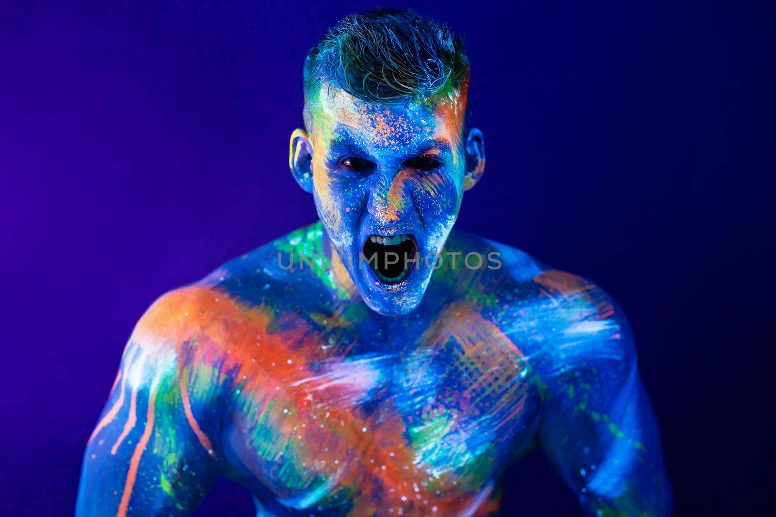 Young man shouts. Fluorescent paint on face and muscular torso, in studio shot with UV light. by StudioPeace