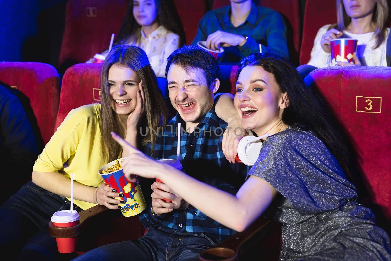 Two young girls and a guy watching a comedy in a cinema. Young friends watching movie in cinema. Group of people in theater with popcorn and drinks
