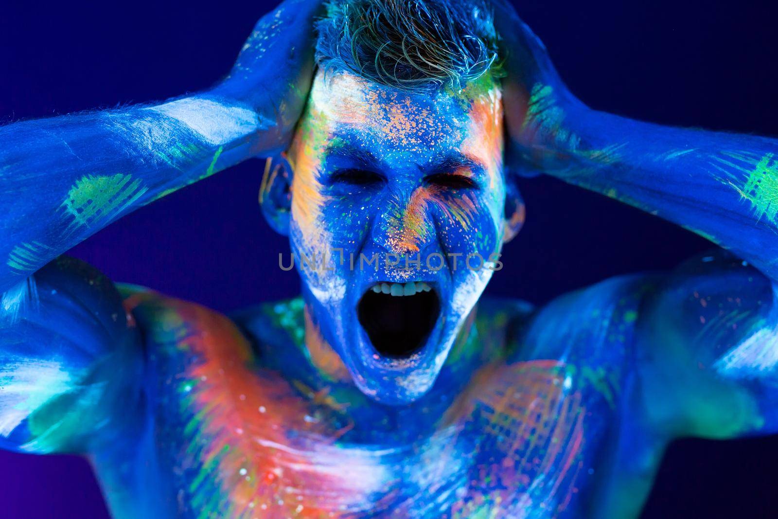 Young man shouts. Fluorescent paint on face and muscular torso, in studio shot with UV light