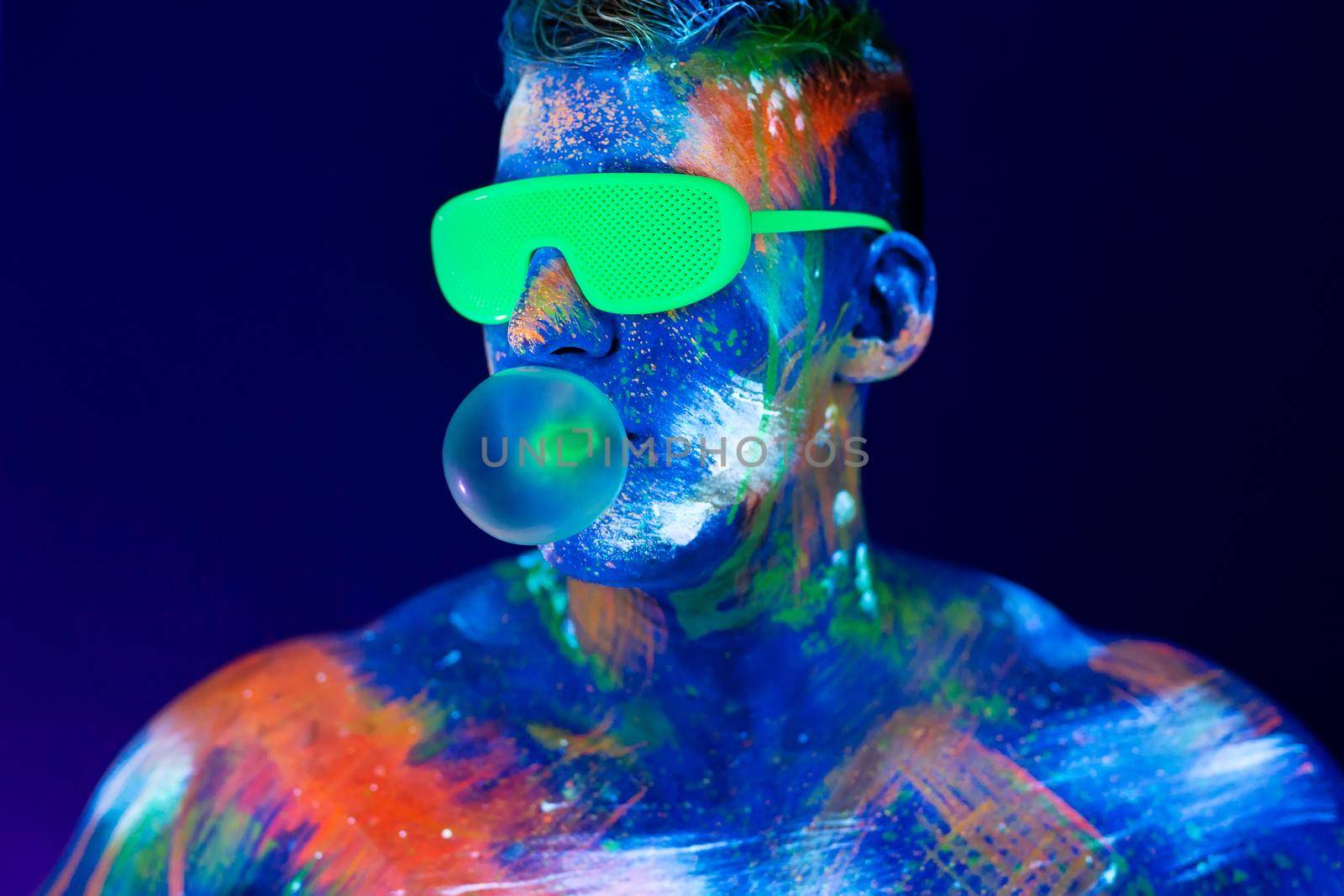 Young man chews gum and inflates a balloon. Fluorescent paint on face in studio with UV light. by StudioPeace