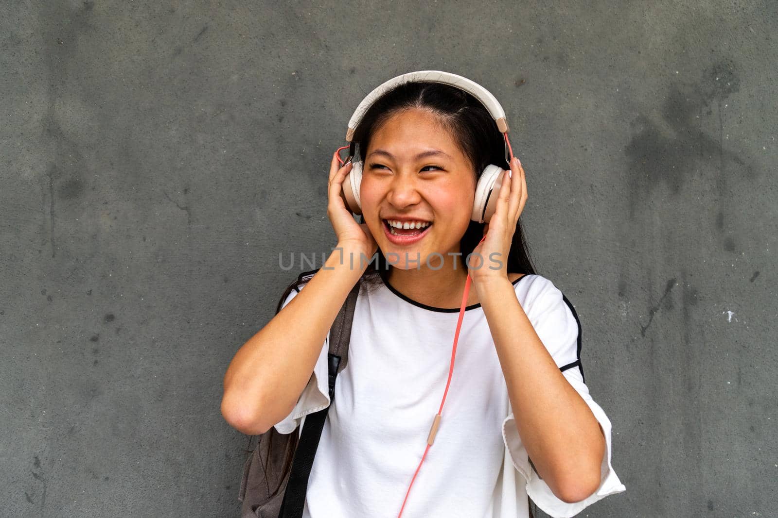 Happy teen asian girl listening to music with headphones on dark concrete background. by Hoverstock