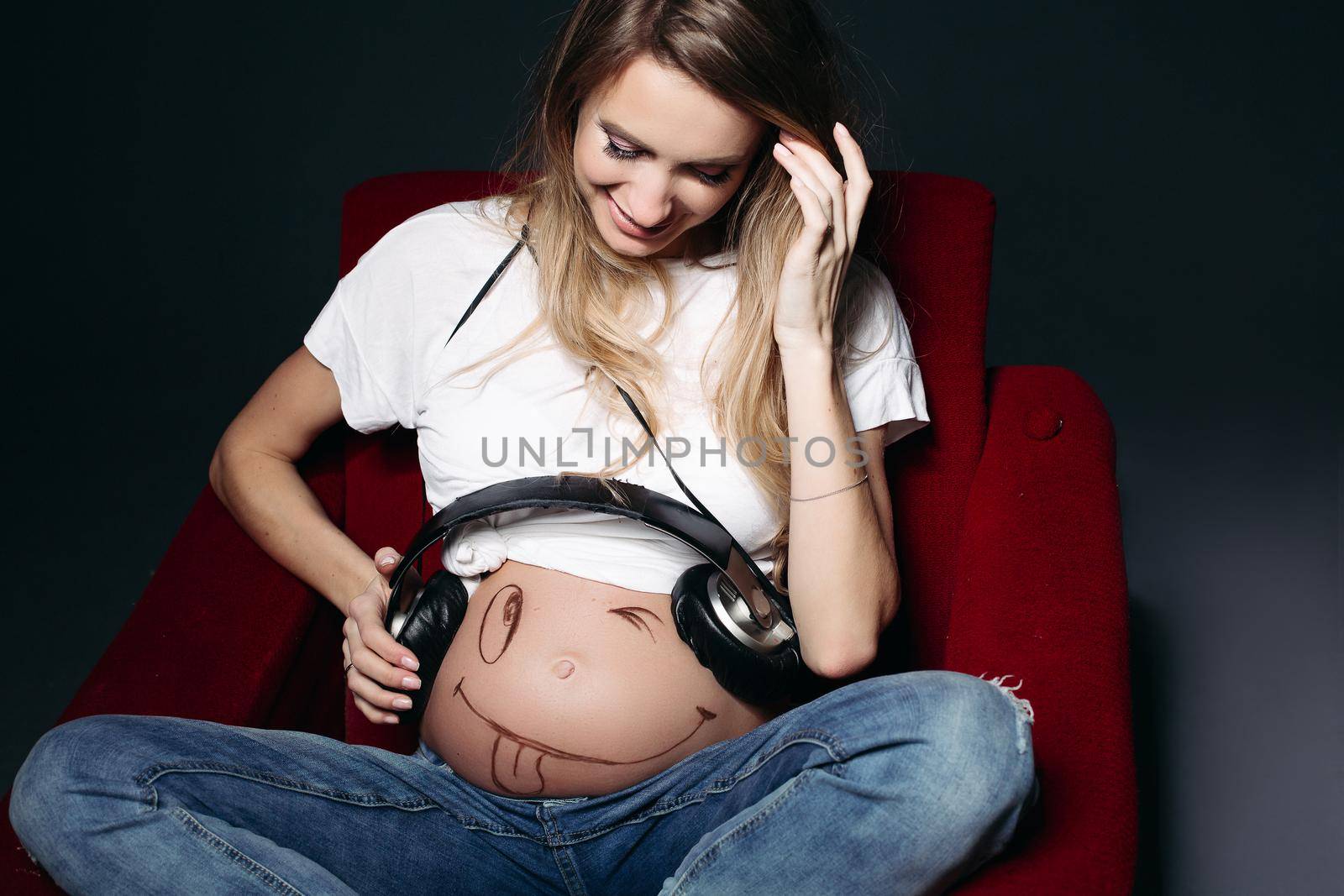 Happy and smiling pregnant female wearing white t shirt and jeans with big earphones on tummy. Expectant mother painting funny smile on her naked belly. Concept of maternety and health care.