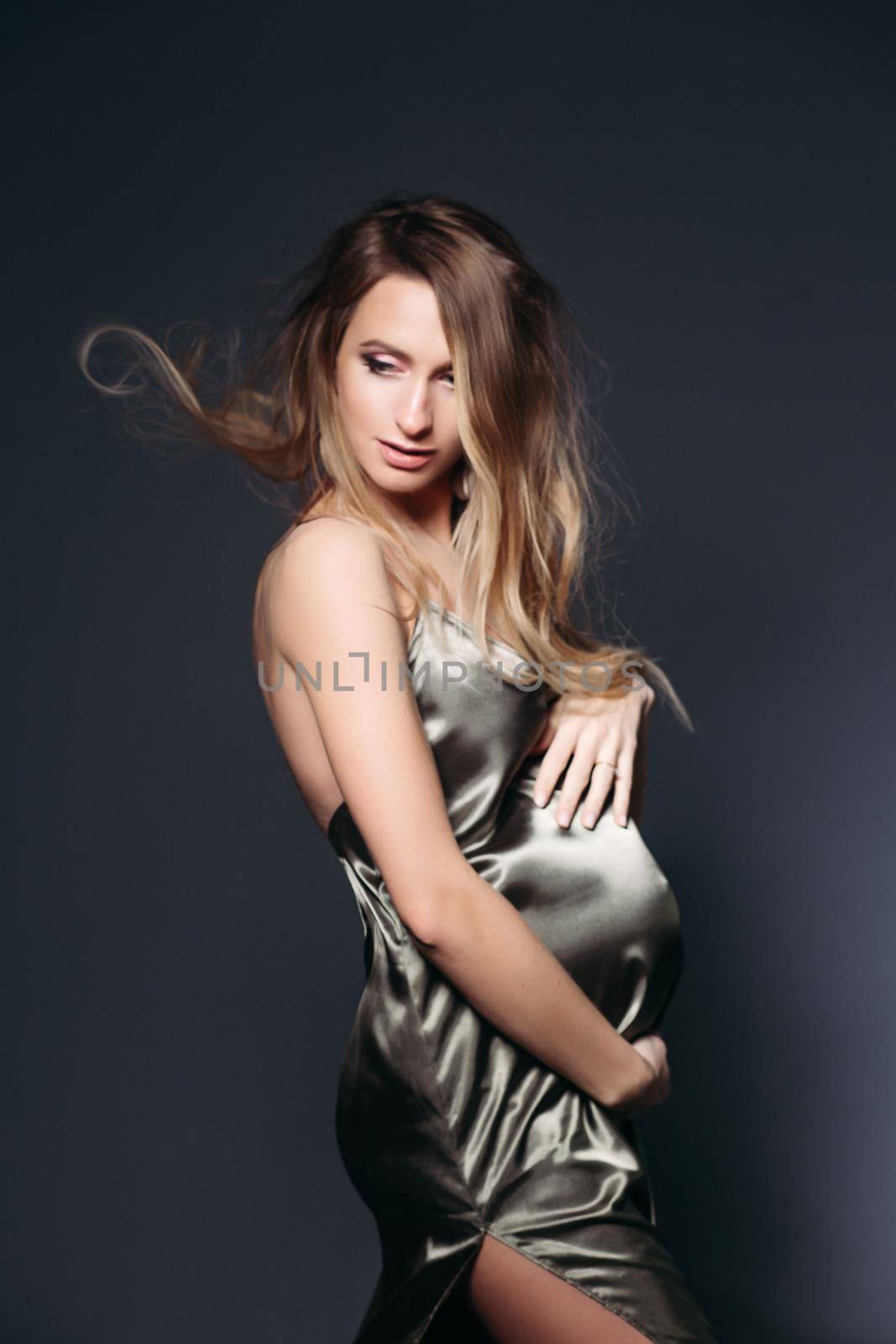 Blondie pegnant female, waiting for a baby, posing at dark studio in gray silk dress with hand up, looking at camera. Elegant future mother expacting her baby. Expextancy of motherhood. Stylish look.
