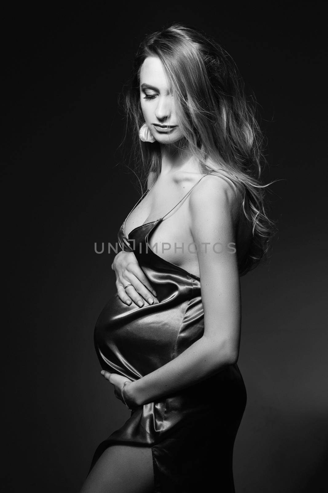 Monochrome portrait of gorgeous and beautiful pregnant woman in sexy silk dress, embracing belly. Fashionable mom posing at dark studio, looking at camera. Concept of pragnancy fashion. Windy effect.