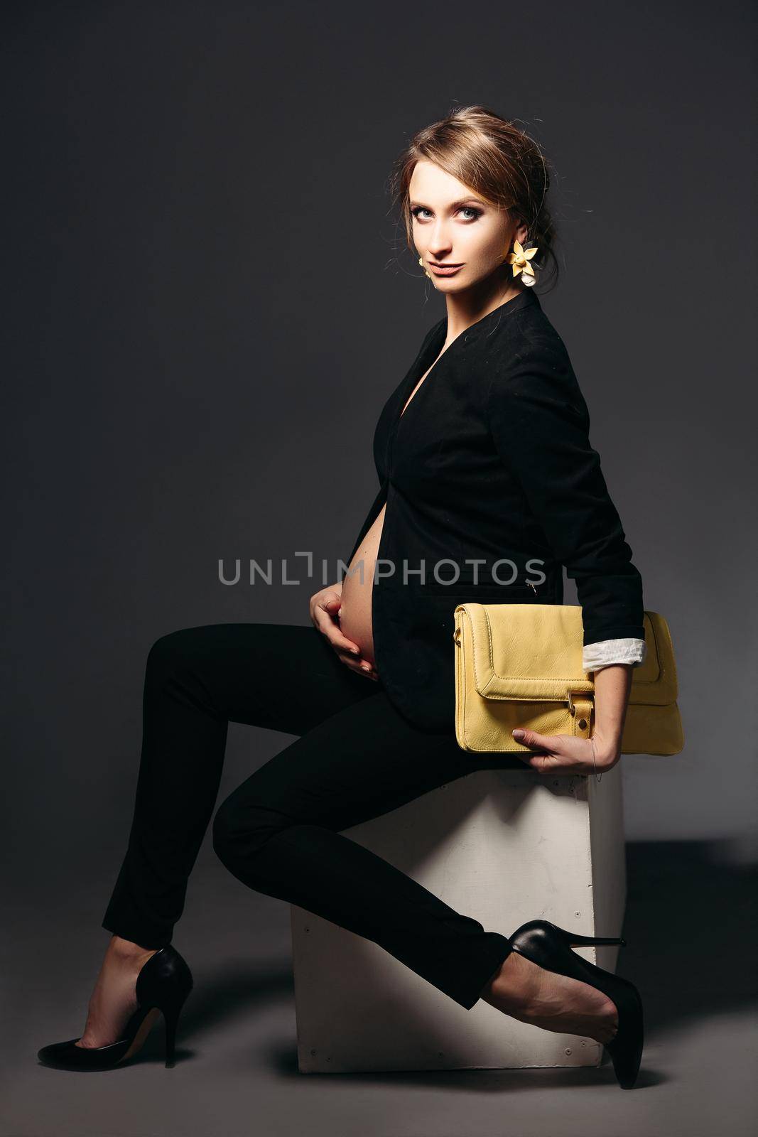 Portrait of elegant pregnant woman wearing in black suit with pregnant bell looking under jacket. Brunette girl holding yellow handbag, sitting against black studio background and looking at side.