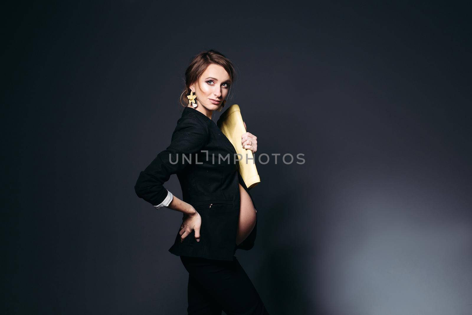 Portrait of elegant fashionable pregnant woman with long earrings, wearing in modern black suit, with yellow bag in one hand and holding another hand on her waist. Pregnant bell looking under jacket.