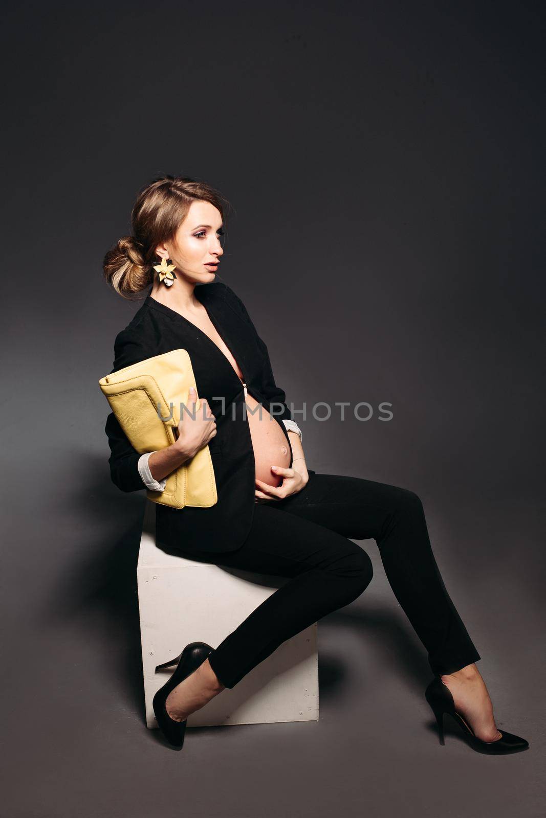 Portrait of elegant pregnant woman wearing in black suit with pregnant bell looking under jacket. Brunette girl holding yellow handbag, sitting against black studio background and looking at side.