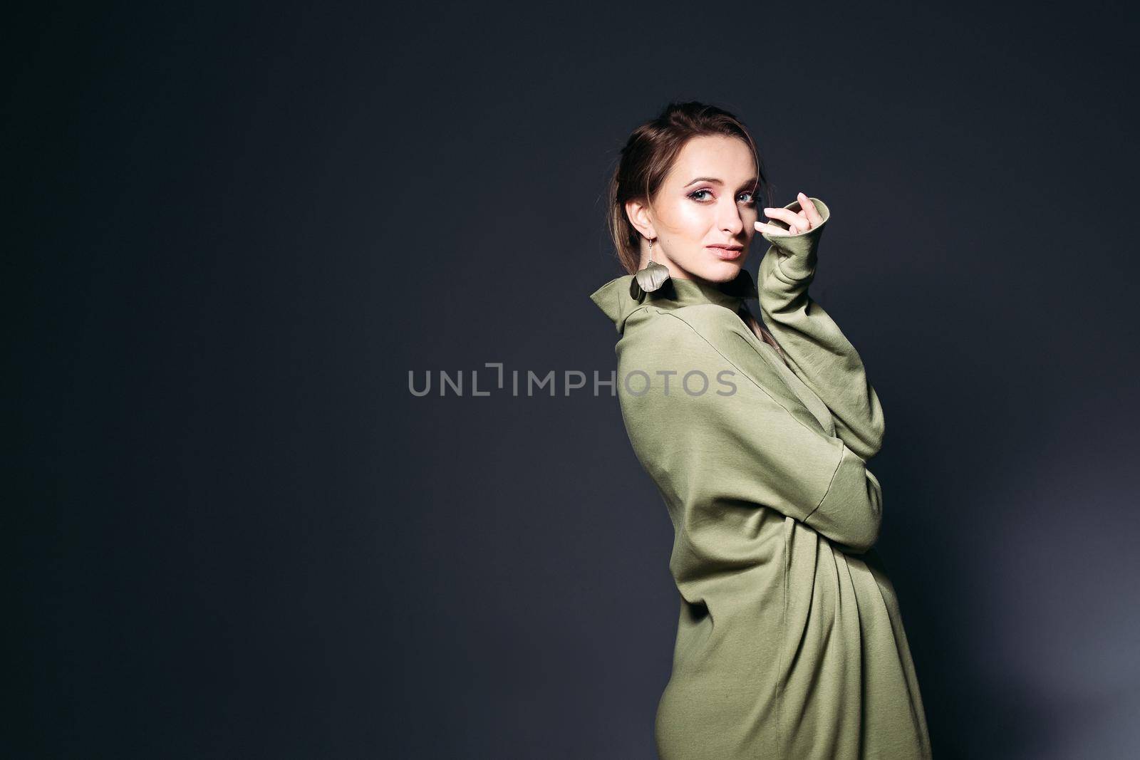 Elegant and fashionable pregnant woman posing at dark studio, sensuality touching face by hands. Stylish woman wearing in khaki dress with pregnant belly, looking at camera. Concept of pregnancy.