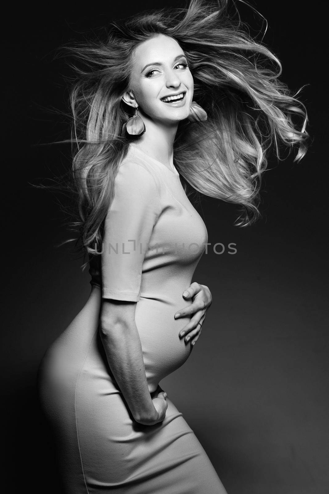 Monochrome portrait of beautiful, happy pregnant woman in dress, embracing belly and smiling. Positivity and stylish female posing, looking at camera. Concept of pragnancy fashion. Windy effect.