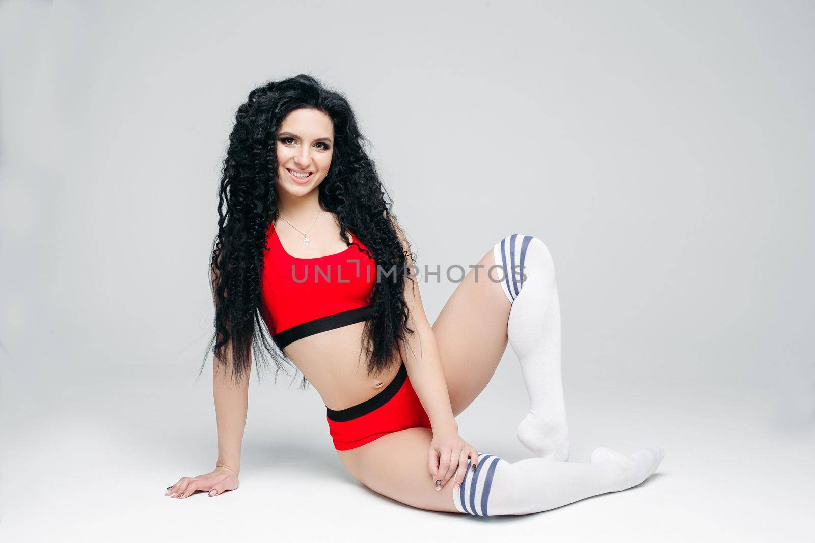 Full lenght portait of sexy dark haired girl wearing in red sports wear and white leggings lying on floor. Woman posing at white studio background, smiling at camera. Concept of sport, fitness, dance.