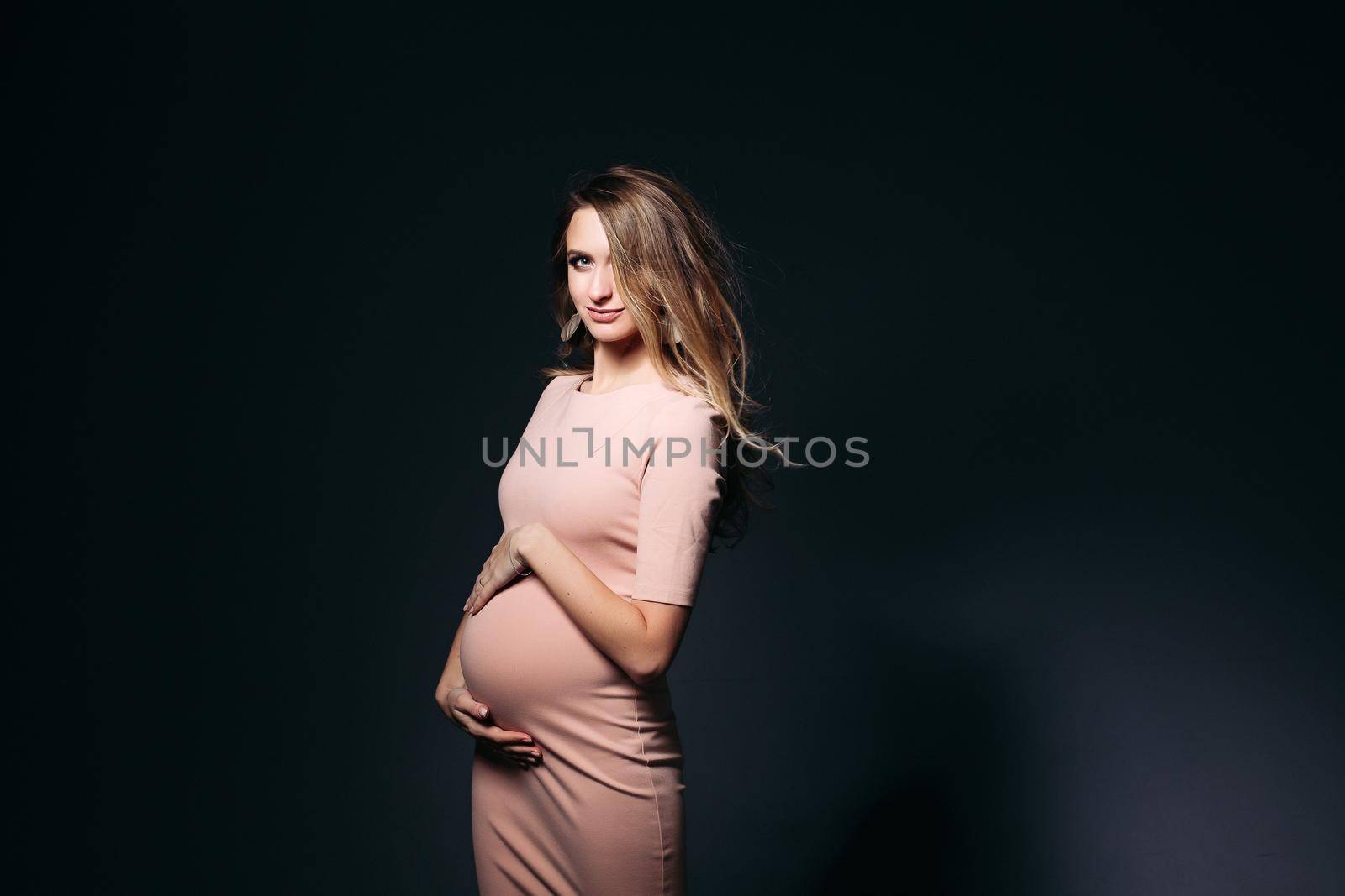 Windy effect on studio portrait of beautiful pregnant woman in pink dress, embracing belly. Elegant and stylish female posing, looking at side. Concept of pragnancy fashion and look.