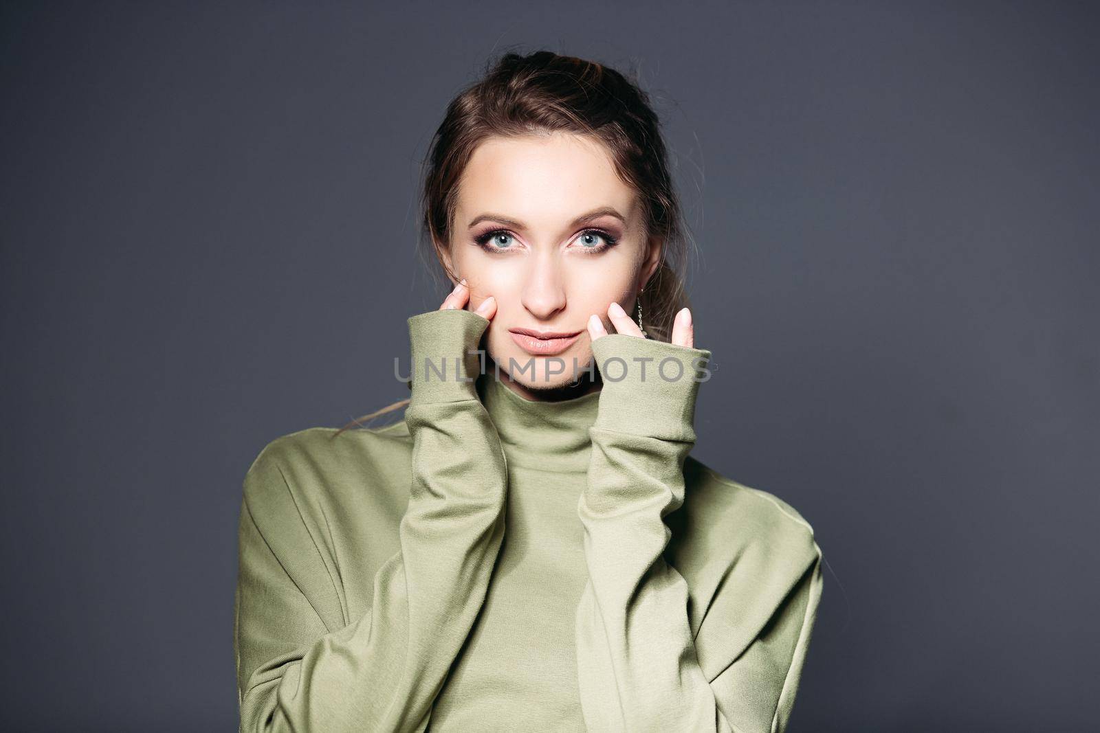Close up of young brunette woman with big blue eyes, plump lips, wearing in khaki dress posing at studio, touching skin. Woman with perfect make up touching face by fingers, looking at camera.