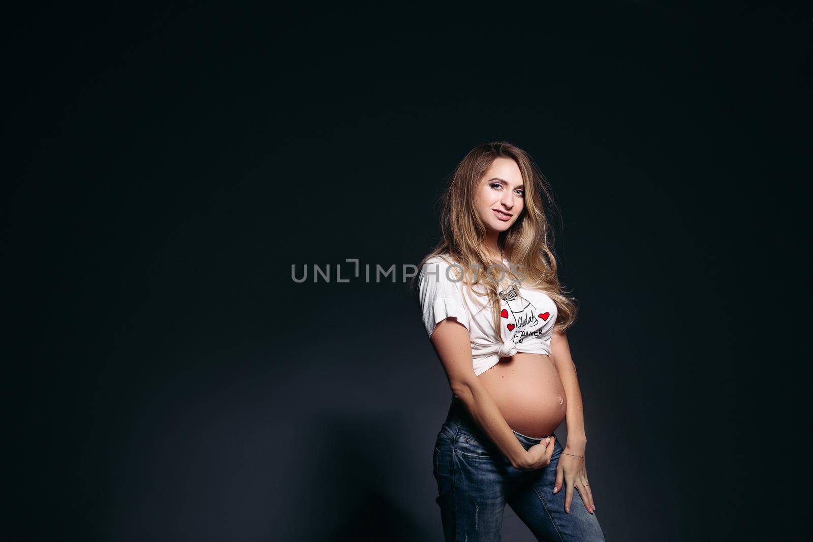 Front view of pretty and fashionable pregnant woman wearing in jeans and white t shirt embracing stomach. Future mother smiling at camera,holding hands on her naked tummy. Concept of pregnant fashion.