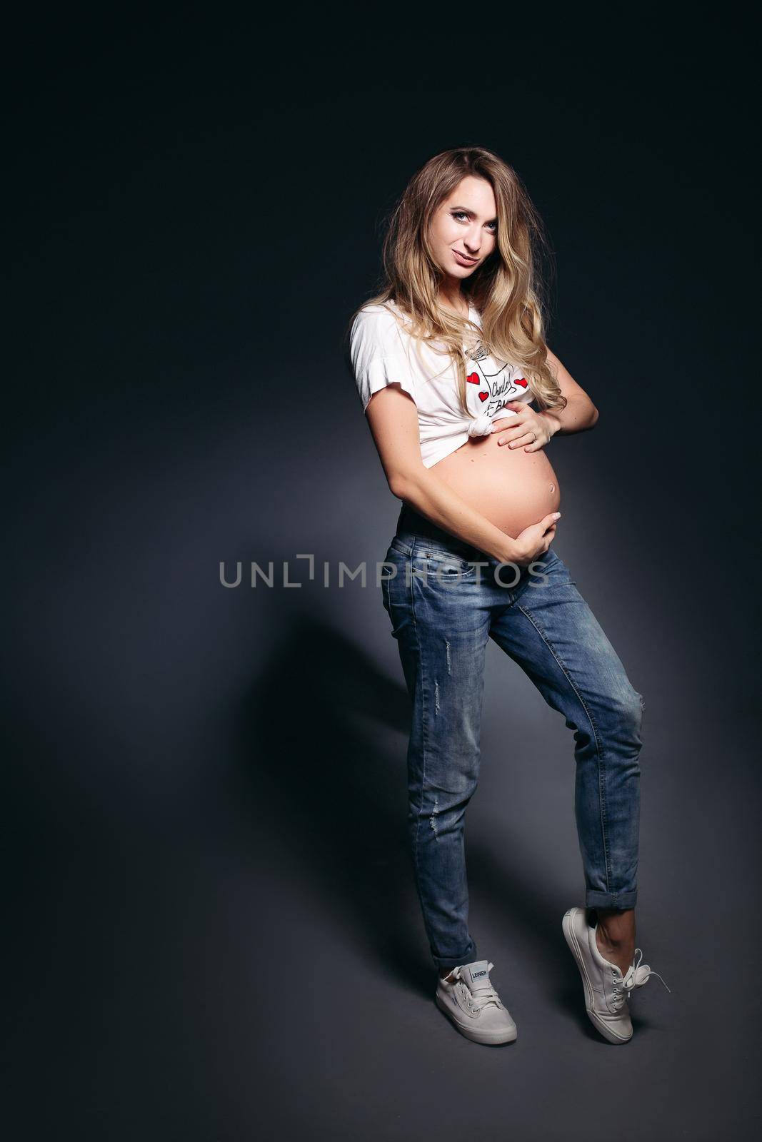 Front view of pretty and fashionable pregnant woman wearing in jeans and white t shirt embracing stomach. Future mother smiling at camera,holding hands on her naked tummy. Concept of pregnant fashion.