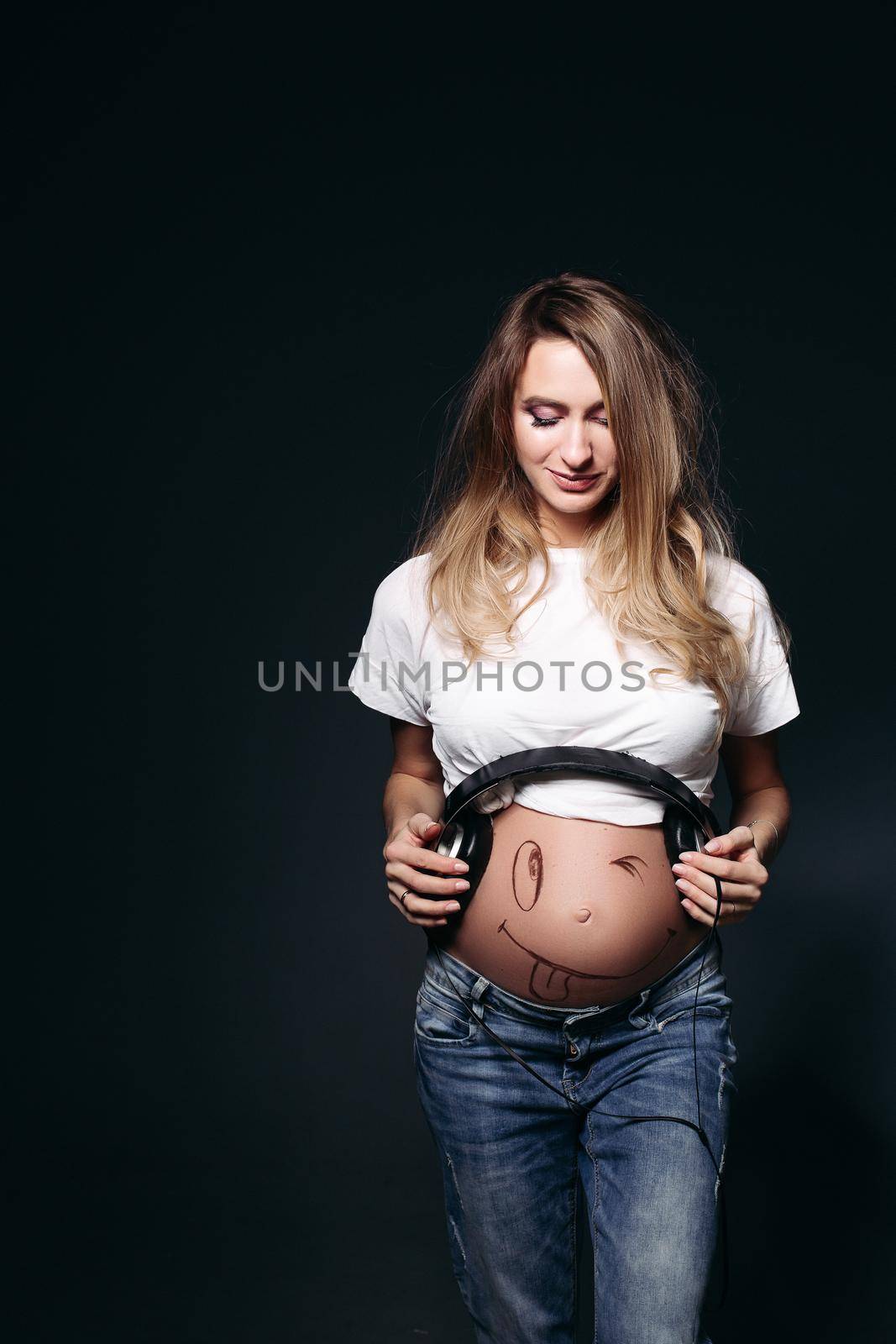 Happy and smiling pregnant female wearing white t shirt and jeans with big earphones on tummy. Expectant mother painting funny smile on her naked belly. Concept of maternety and health care.