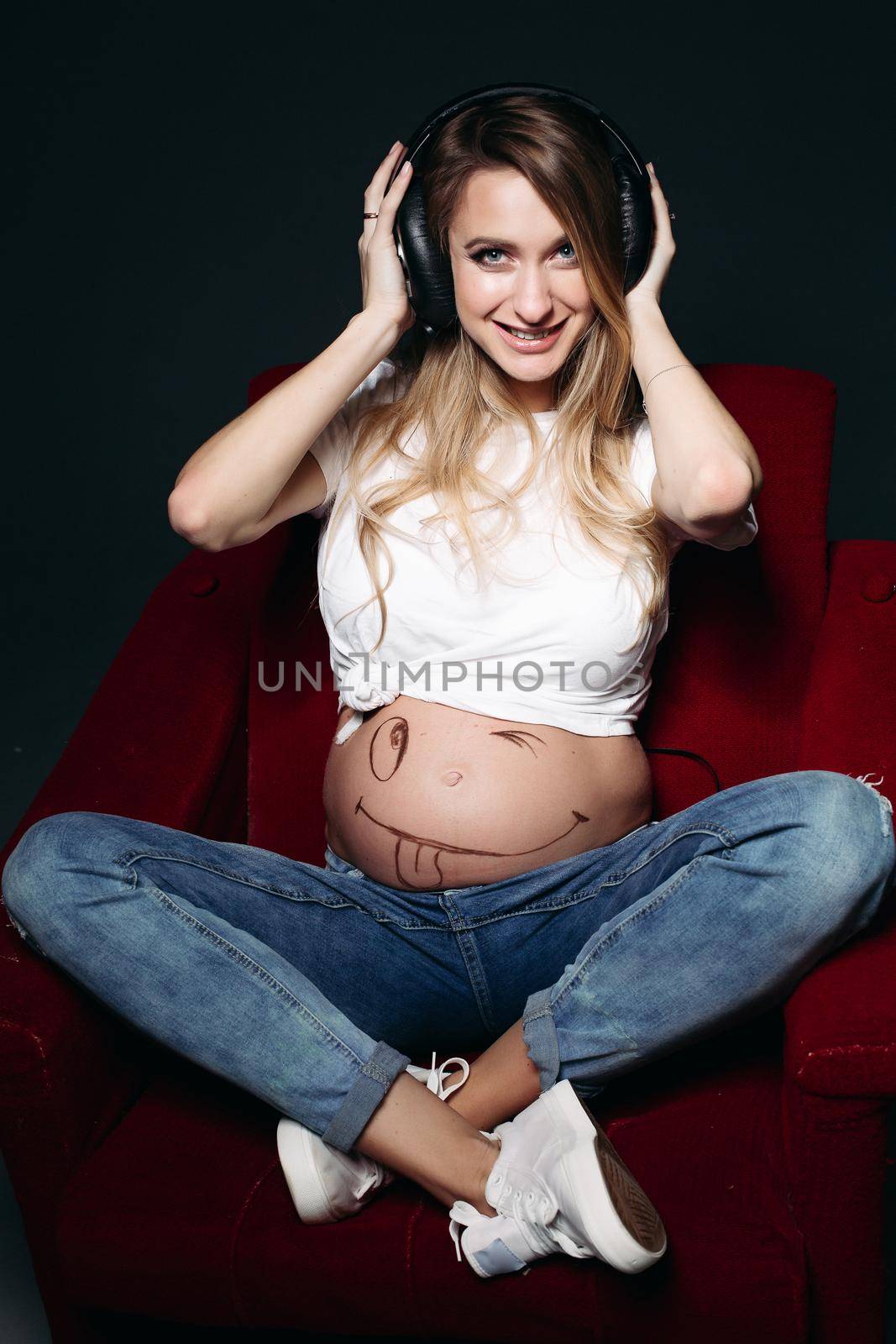 Happy and smiling pregnant female wearing white t shirt and jeans with big earphones on tummy. Expectant mother painting funny smile on her naked belly. Concept of maternety and health care.