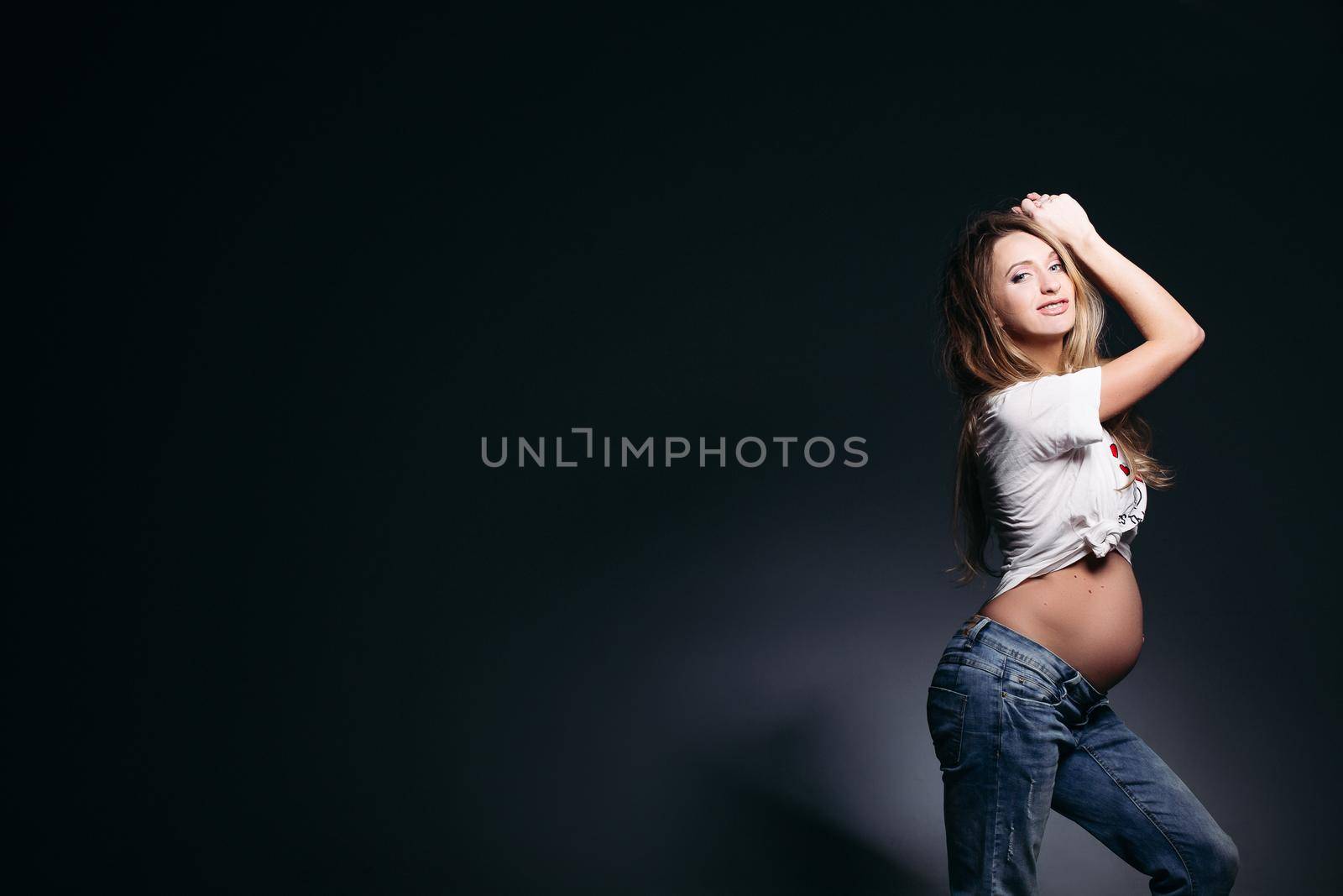 Side view of beautiful and fashionable pregnant woman in jeans and t shirt posing at dark studio background. Stylish and smiling mom posing with naked tummy, holding hand up, looking at camera.