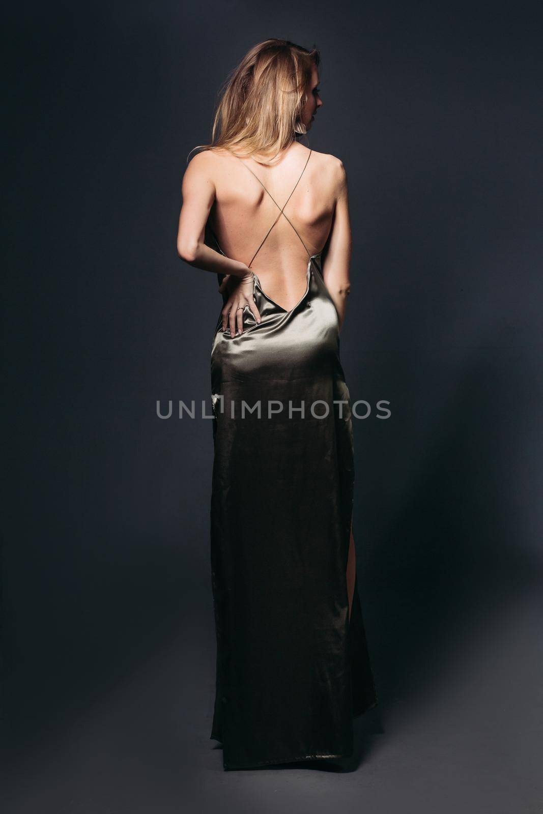Back view of incognito elegant and sexy blondie with nude shoulder and perfect hairstyle, posing at sexy long dress. Fashionable look at studio against gray background. Shopping, sale, fashion.