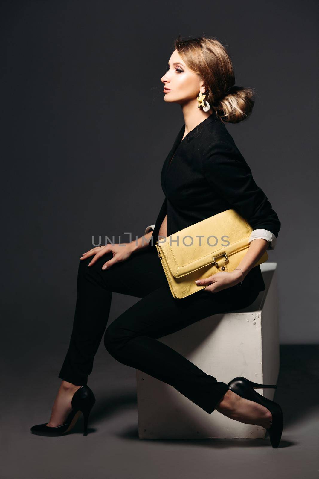 Portrait of elegant pregnant woman wearing in black suit with pregnant bell looking under jacket. Brunette girl holding yellow handbag, sitting against black studio background and looking at side.