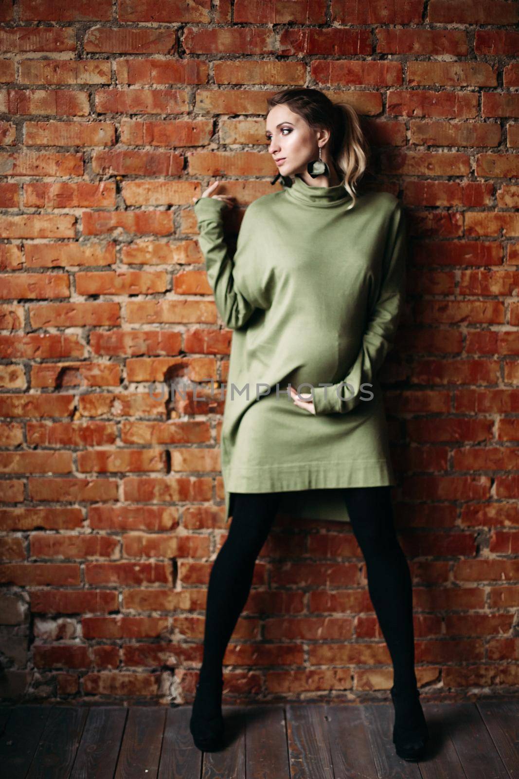 Portrait of happy pregnant brunette girl posing against brick wall. Stylish woman wearing in khaki dress, holding pregnant belly by hands, looking at side. Concept of pregnancy and paternity.