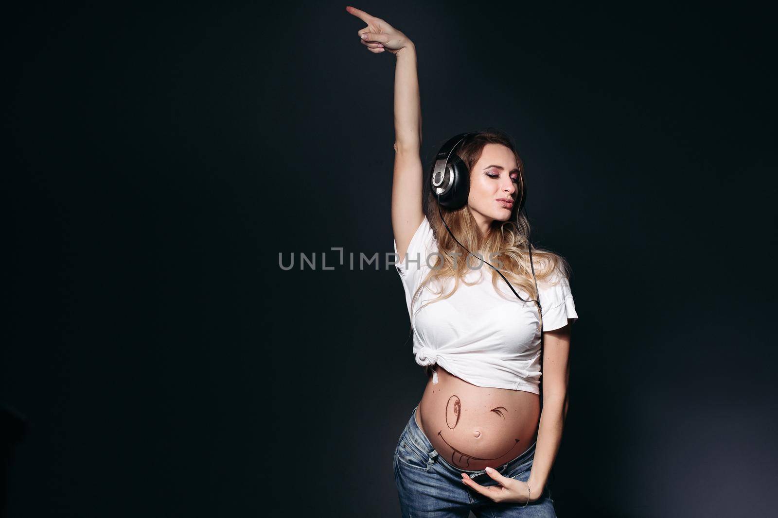 Beautiful pregnant mom dancing, smiling, posing with closed eyes. Expectant mother rising up, listening music by earphones and showing naked tummy. Lady with long hair, wearing in shirt and jeans.