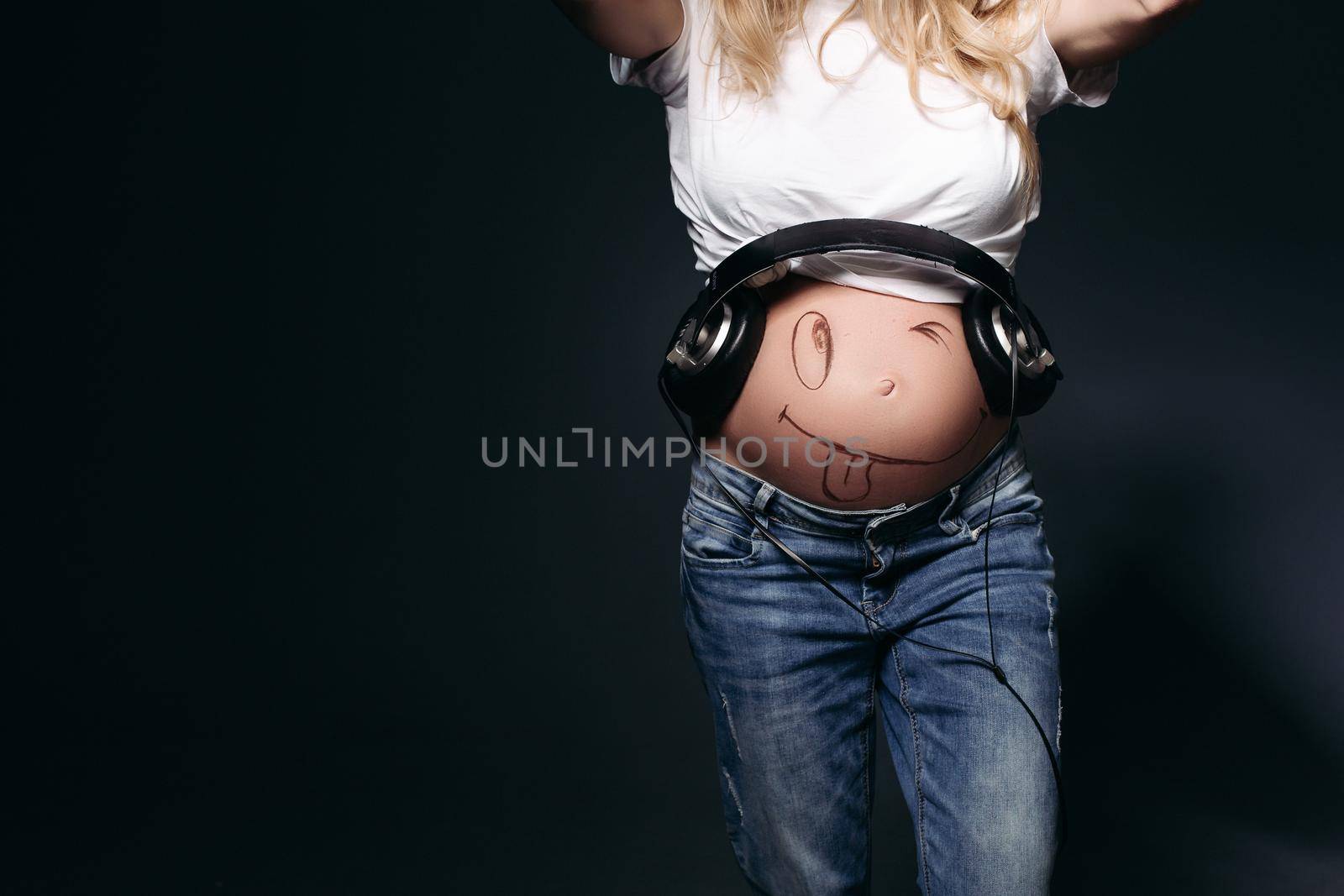 Crop of stylish pregnant incognito mom wearing white t-shirt and jeans with big earphones on tummy. Expectant mother painting funny smile on her naked belly. Black studio background.