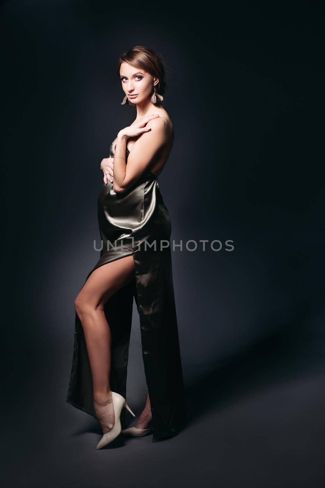 Studio portrait of sensual and beautiful pregnant woman in sexy silk dress, embracing belly. Elegant and stylish female posing, looking at side. Concept of pragnancy fashion and look.