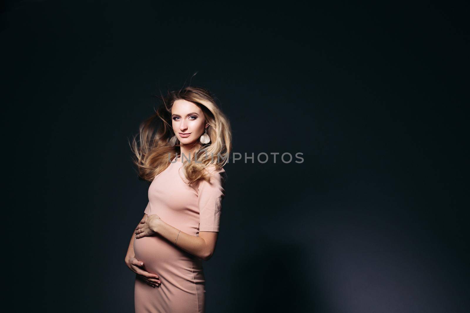 Windy effect on studio portrait of beautiful pregnant woman in pink dress, embracing belly. Elegant and stylish female posing, looking at side. Concept of pragnancy fashion and look.