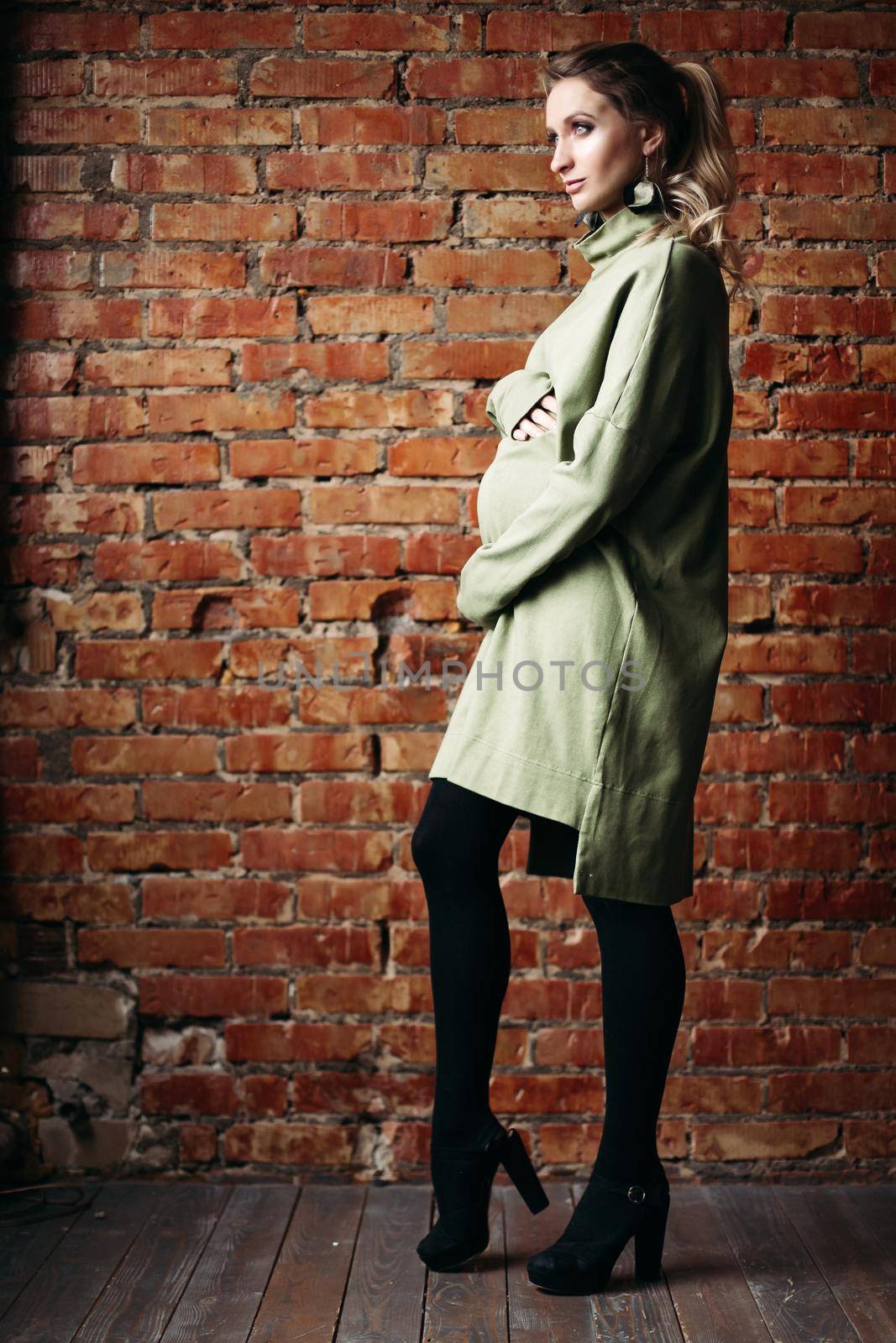 Portrait of happy pregnant brunette girl posing against brick wall. Stylish woman wearing in khaki dress, holding pregnant belly by hands, looking at side. Concept of pregnancy and paternity.