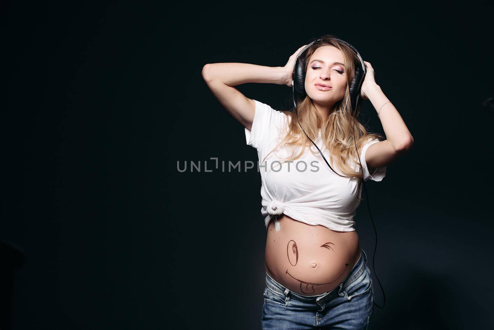 Portrait of beautiful pregnant woman with long hair dancing, smiling with closed eyes. Expectant mother with naked tummy listening music and holding big earphones by hands. Stylish mom wearing in jeans.