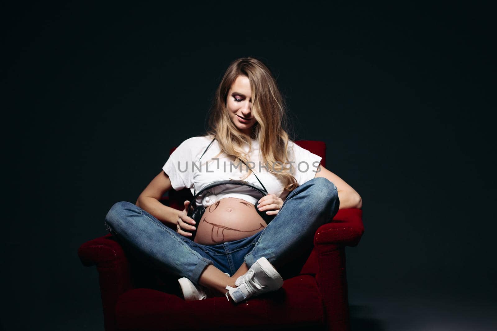 Front view of adorable expectant mother sitiing on red chair and holding earphones on her tummy with painted smile. Lady wearing blue jeans and white tee shirt. Concept of maternity. Black background.