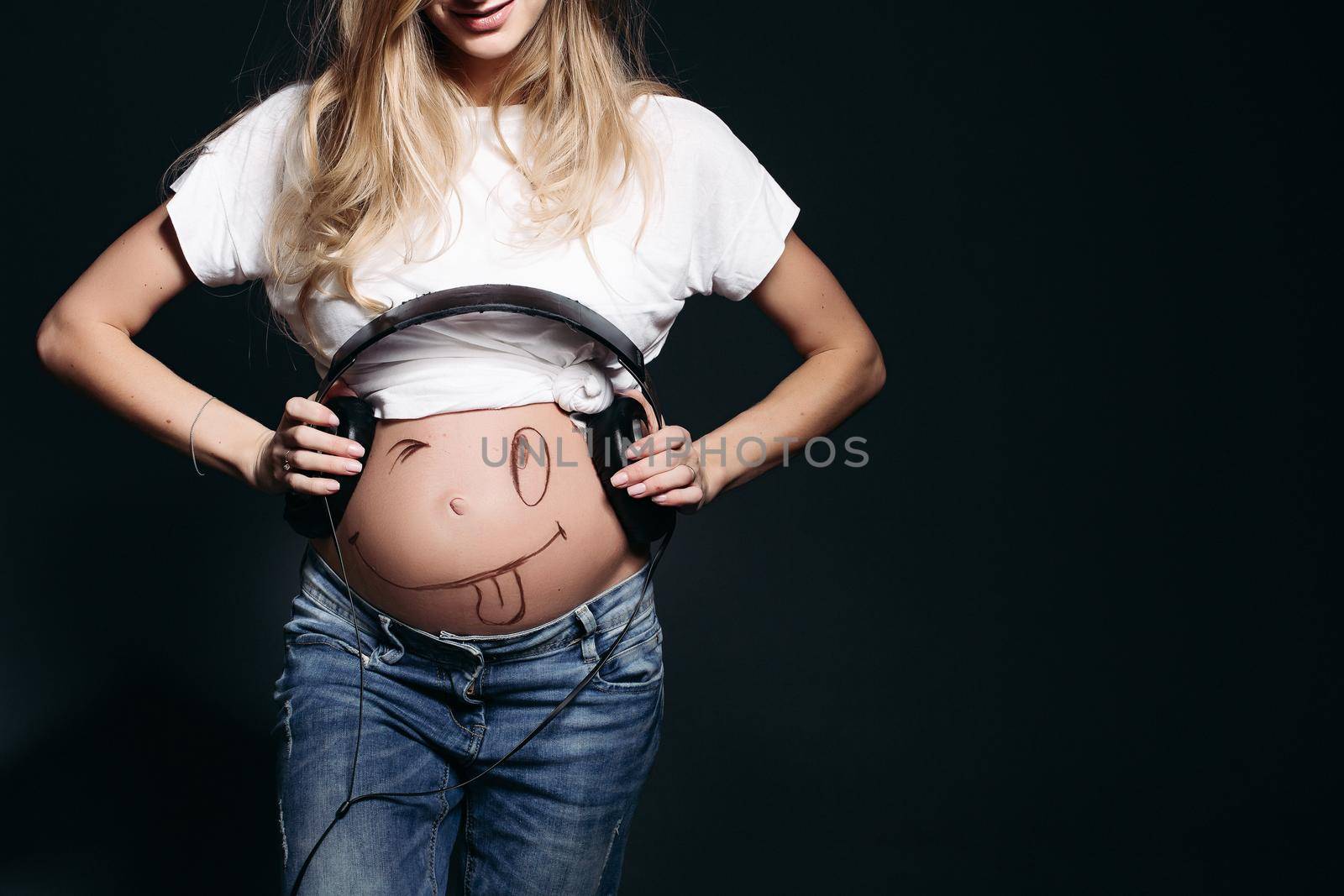 Happy and smiling pregnant female wearing white t shirt and jeans with big earphones on tummy. Expectant mother painting funny smile on her naked belly. Concept of maternety and health care.
