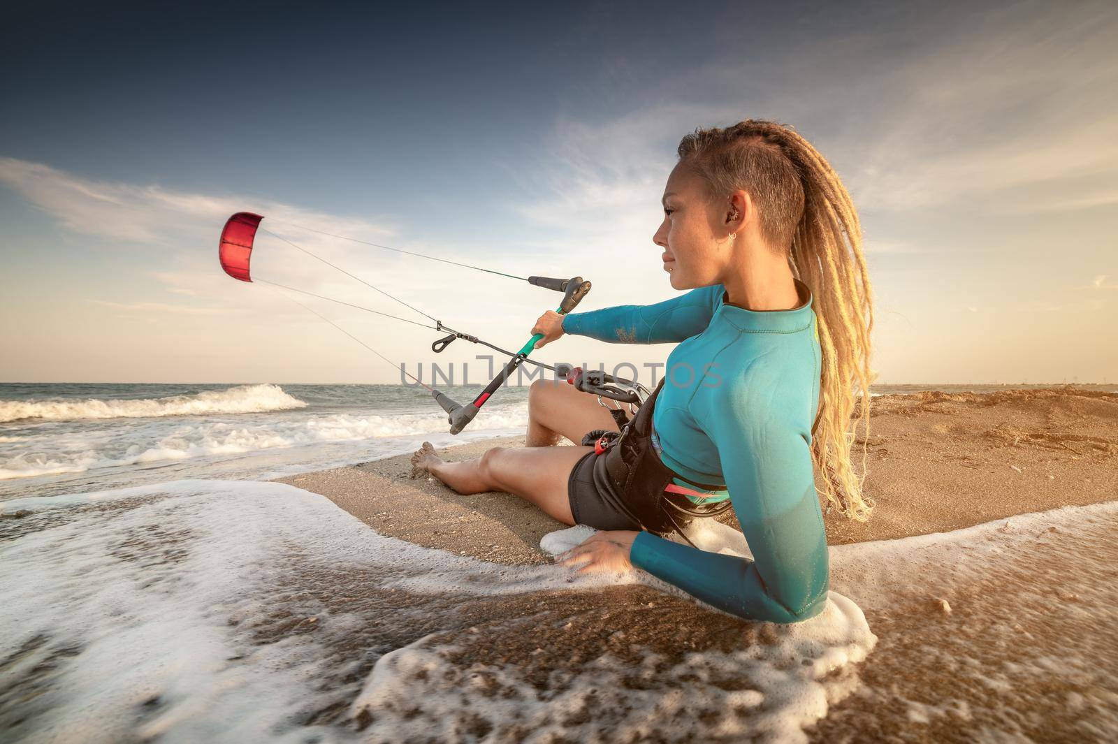 Attractive caucasian woman kitesurfer in a neoprene wetsuit and with dreadlocks on her head is resting lying on a sandy beach on the shore holding her kite in the wind.