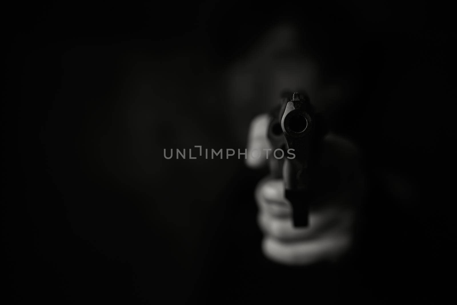Threat of firearms. Muzzle of pistol in person's hand is pointed at camera. Man criminal holds revolver on dark background. Attack or defense. Black and white photo.