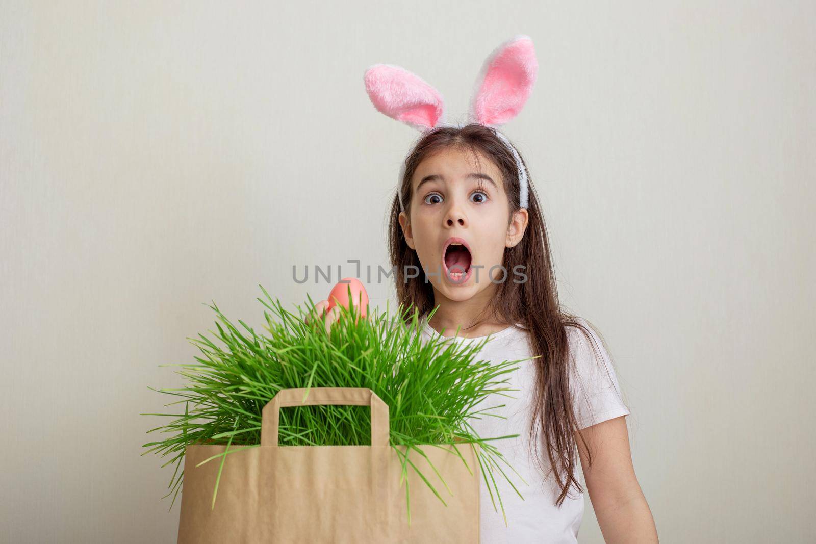 A surprised girl in pink bunny ears hold pink egg from a paper bag with grass.Copy space
