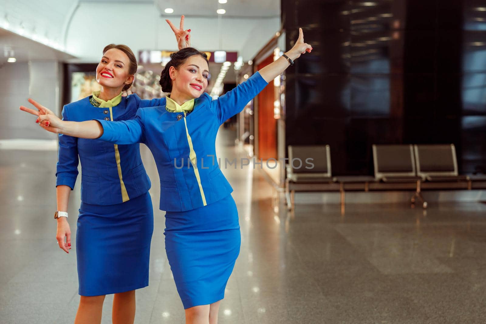 Joyful female flight attendants in air hostess uniform doing peace hand gesture and smiling while standing in airport terminal