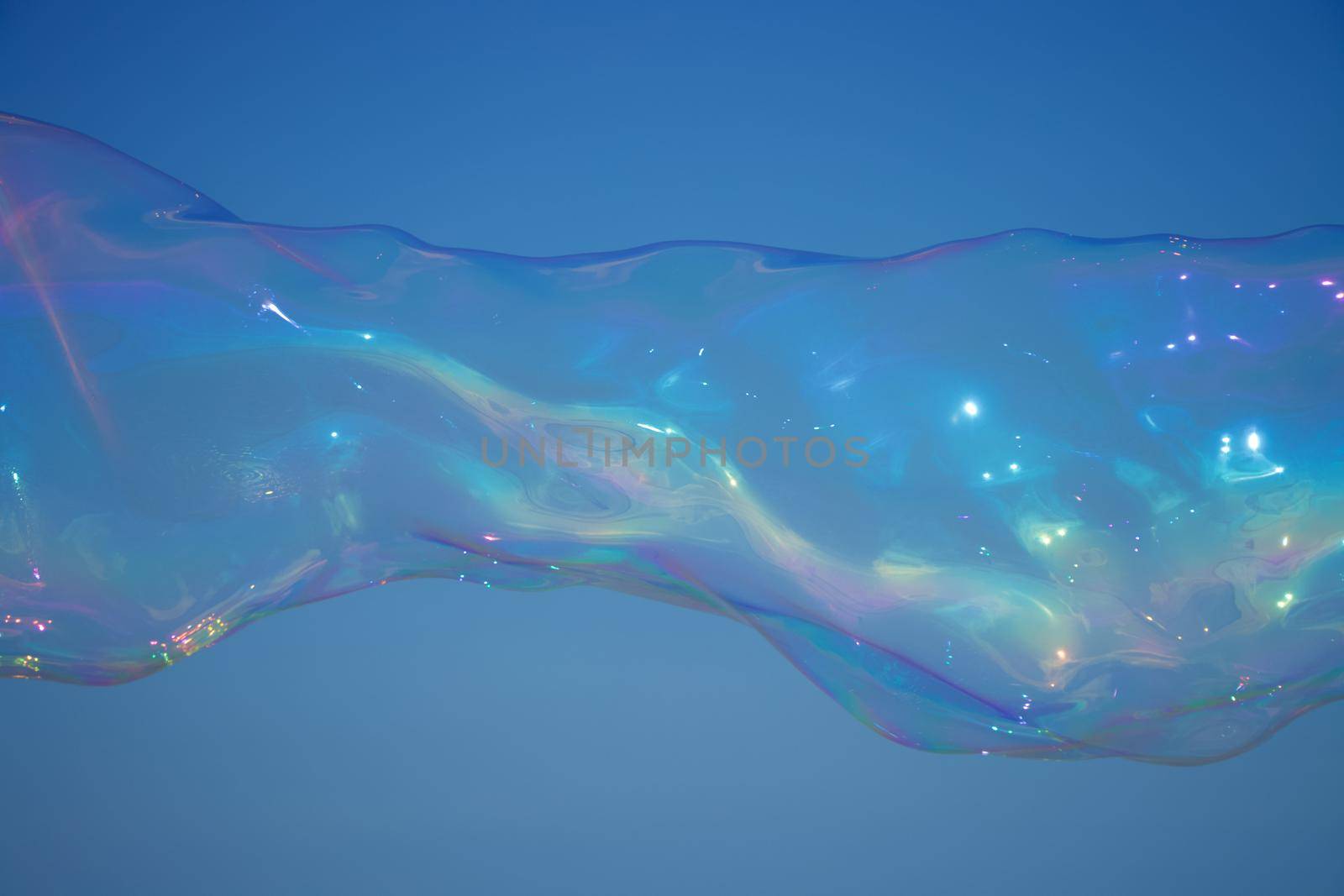 Long, iridescent soap bubble reflecting rainbow colours against a backdrop of a clear blue sky. Captured with Canon EOS 90D on a clear summer's day.