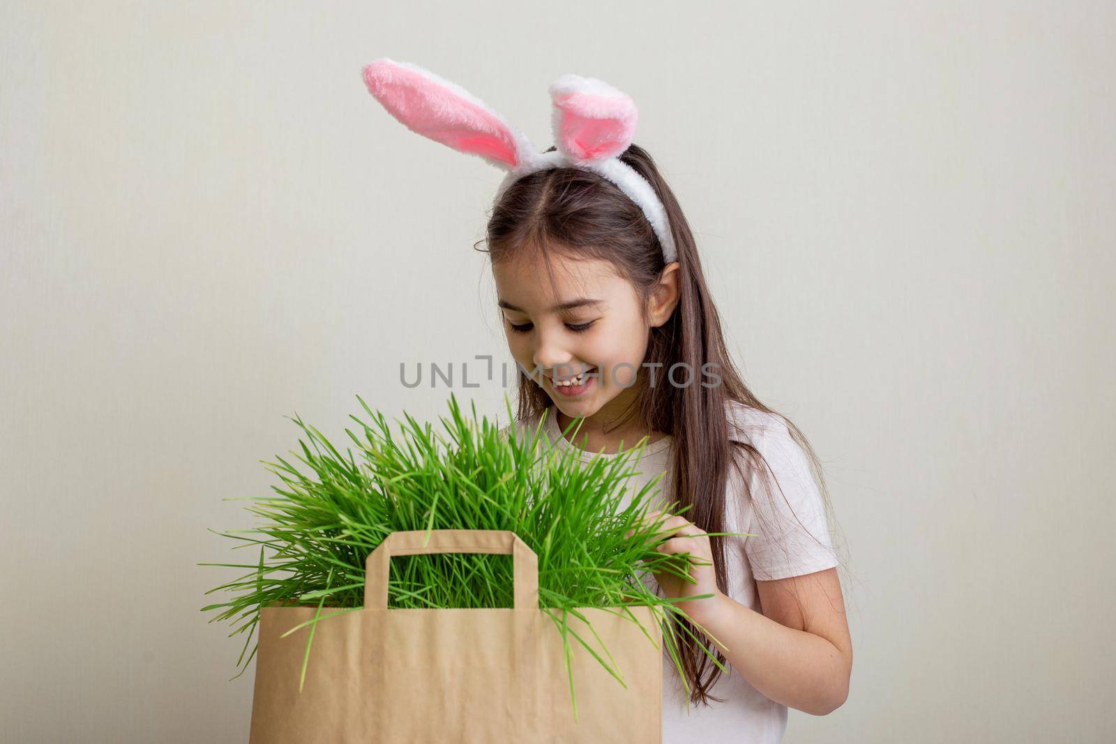 A beautiful little girl in pink bunny ears looks into a paper bag with grass, looking for an Easter egg. copy space
