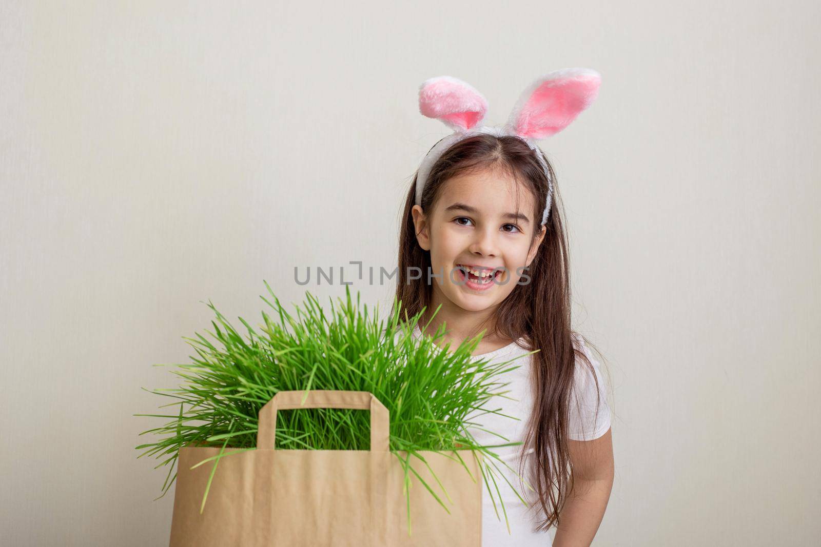 A cute little girl with pink rabbit ears stands next to a paper bag with grass, smiles. Copy space.