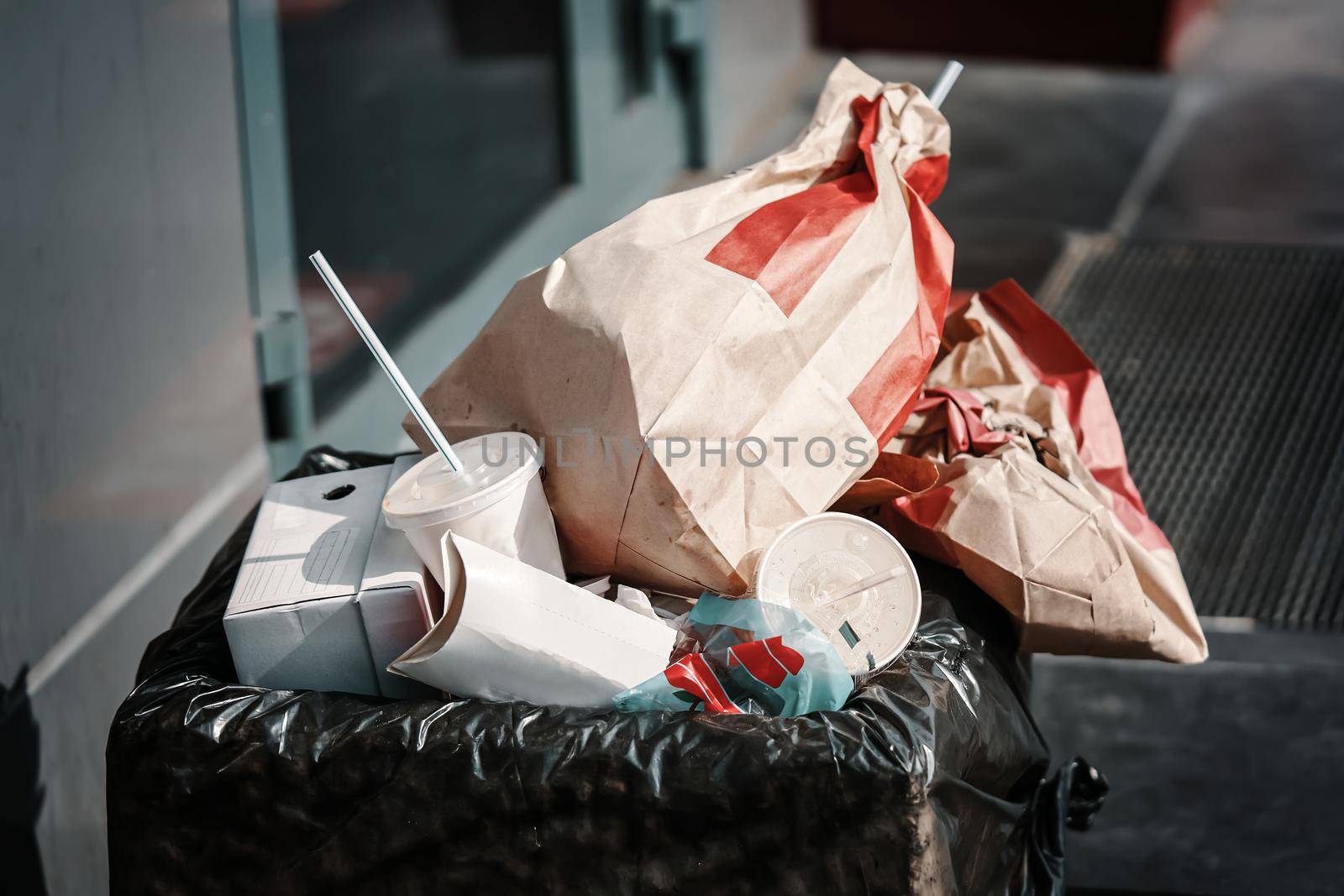 Bunch of paper packets and cups of coffee and other drinks in garbage. Concrete trash can with black bag inside. Dustbin next to fast food outlet. Urban landscape.